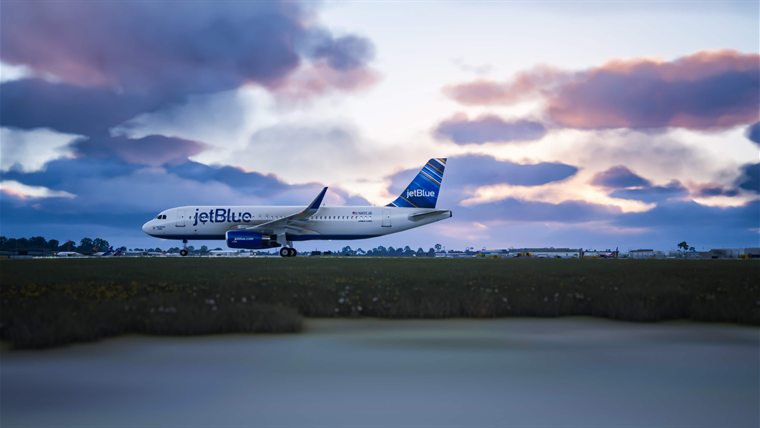 A JetBlue A320 taxiing at dusk under pink and violet clouds, seen across the grass.